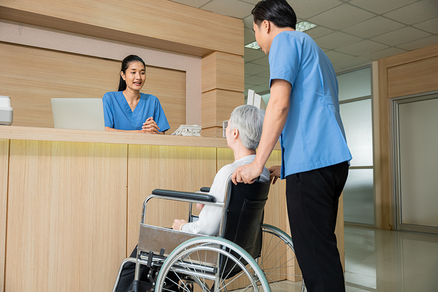 Asian man with grandmother on wheelchair talks to woman in hospital front desk.