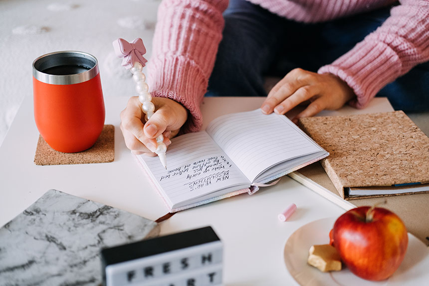 Person writing Chinese New Year's resolutions in a notebook at a desk with a coffee tumbler and apple nearby.