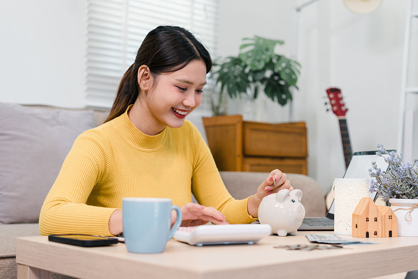 Woman putting coins into a piggy bank while calculating her savings on a calculator.