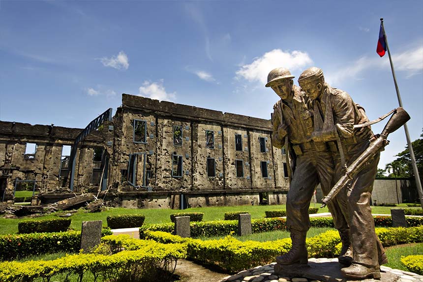 War memorial statue of two soldiers at Corregidor Island with ruined barracks in the background