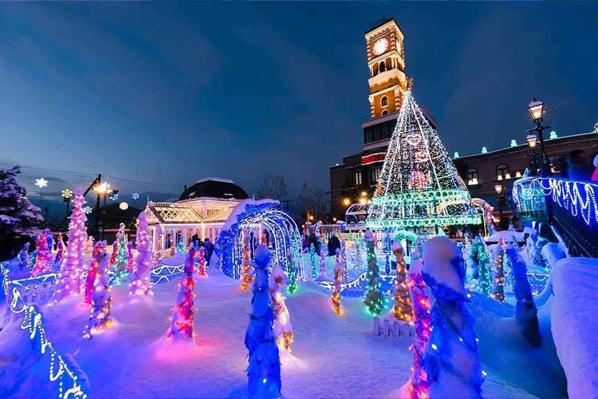 A colorful Christmas tree glows in front of a clock tower, adding holiday cheer to the winter night.
