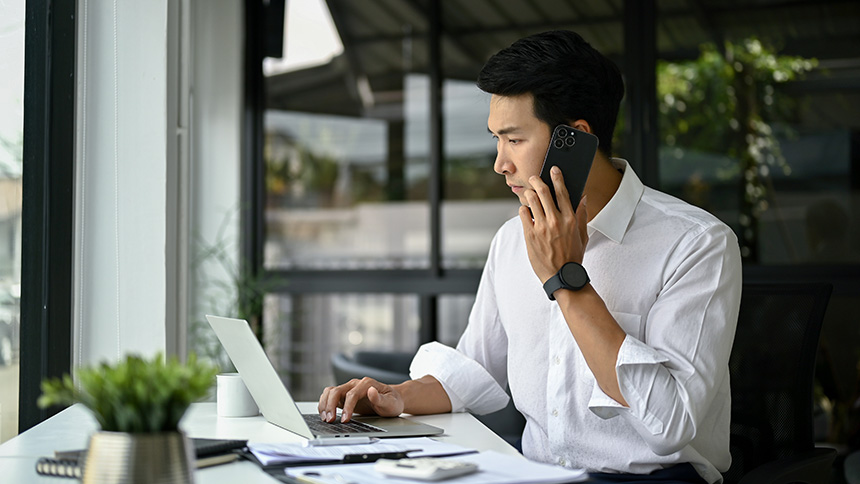 Asian businessman talking to someone on his smartphone while working on his laptop