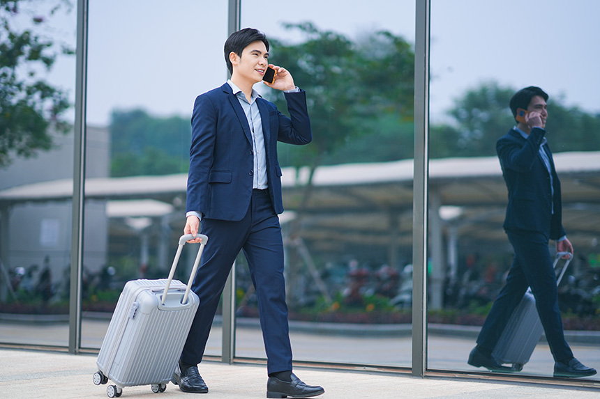 A man in business attire walks with a suitcase, engaged in a phone conversation as he strides forward.