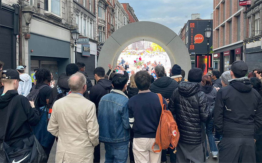 A crowd gathered around the Dublin Portal, watching and interacting with a live video feed.
