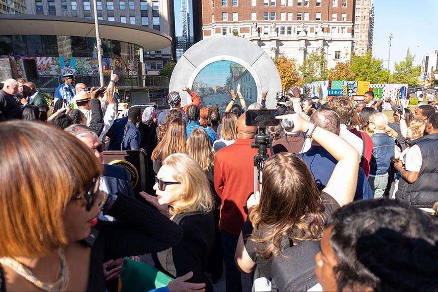 People gathered in the Philadelphia Portal, raising their hands and phones while interacting with the live screen from another city.