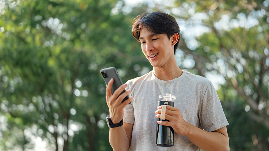 Smiling man outdoors checking his smartphone while holding a reusable water bottle.