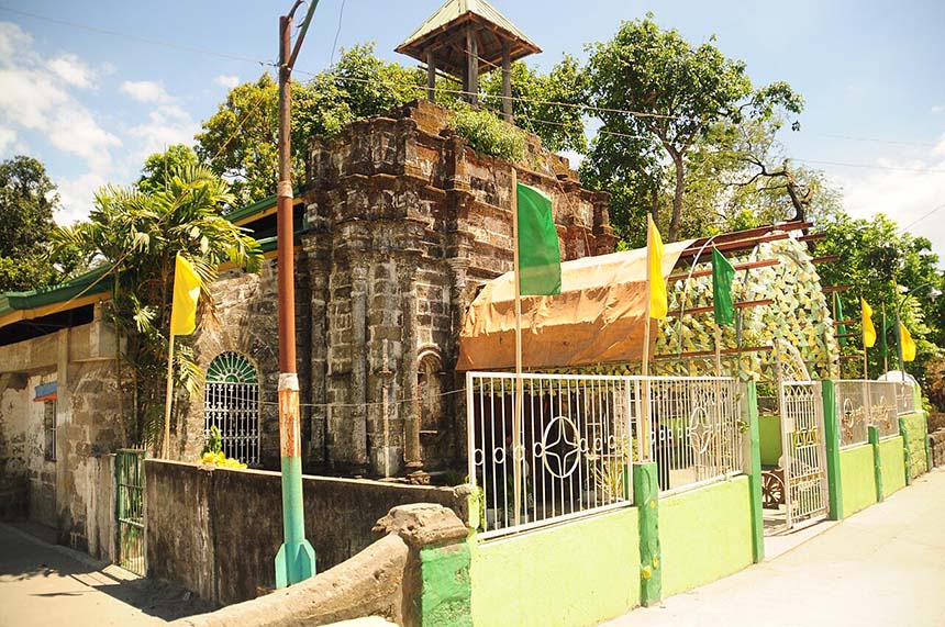 Exterior view of San Jose Matulid Chapel featuring its old stone facade, small bell structure, and surrounding greenery along a quiet roadside.