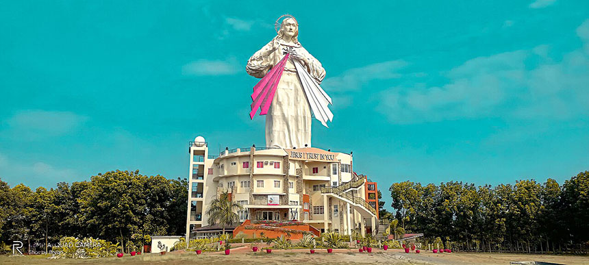 Exterior view of the National Shrine and Parish of the Divine Mercy featuring the towering Divine Mercy statue above the church facade against a clear blue sky.