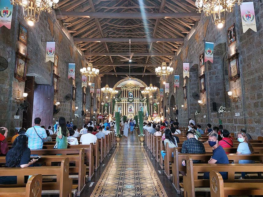 nterior view of Sta. Ursula Parish Church showing stone walls, wooden pews, chandeliers, and worshippers seated facing the altar.