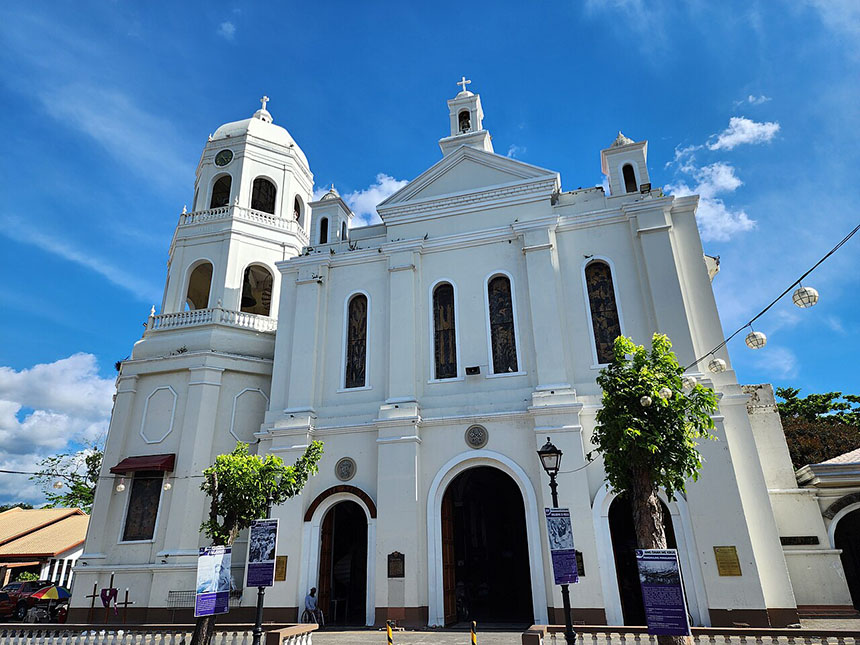 Exterior view of the Minor Basilica of the Immaculate Conception featuring its white colonial facade, bell tower, and arched entrance under a bright blue sky.