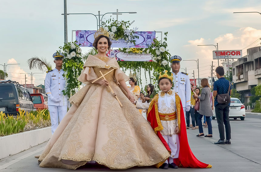 Santacruzan procession with Reyna Elena and child in traditional attire under a floral arch.