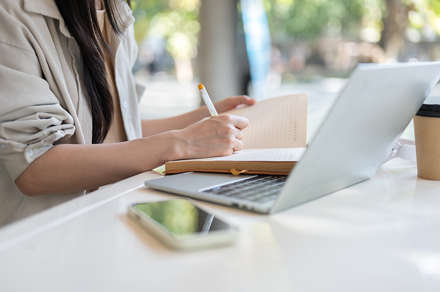 Person working on a laptop while writing in a notebook at home.