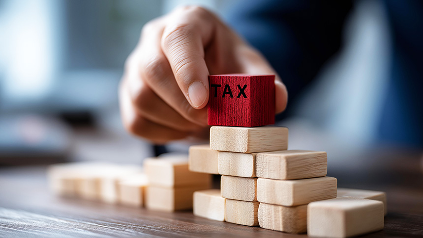 A person placing a red block with the word 'TAX' atop a rising stack of wooden blocks.