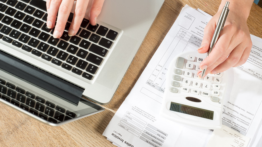 A person typing on a laptop and using a calculator to process business invoices.