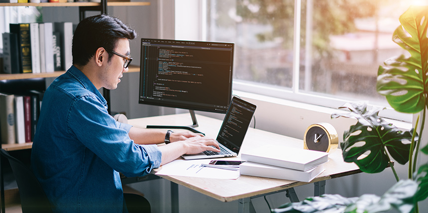 Freelance web developer coding on laptop and monitor at a home office desk.