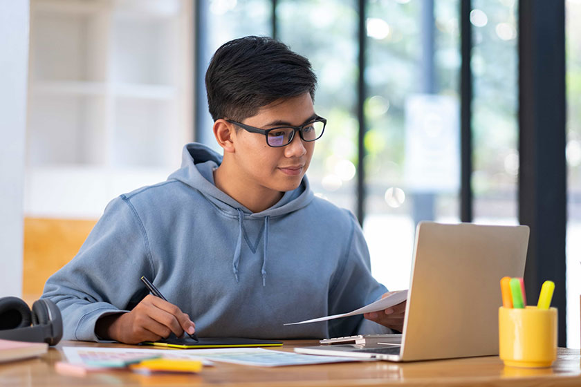 Young man in glasses and a hoodie studying at a desk with a laptop, pen, and papers, in a bright, modern workspace, conveying focus and learning.