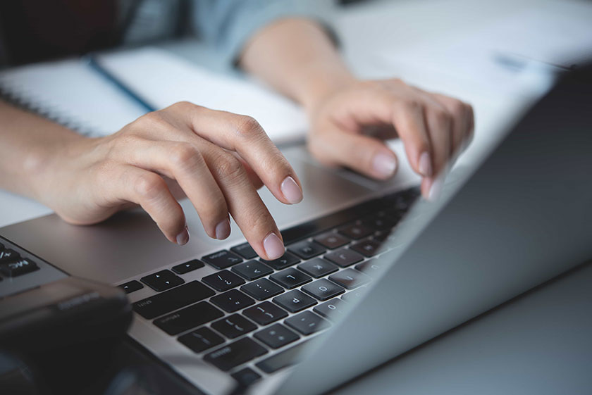 Close-up of hands typing on a laptop keyboard with a notebook and pen in the background, symbolizing productivity and digital work.