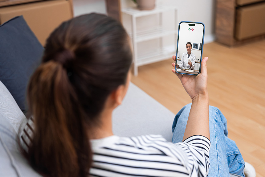 on a couch during a telehealth consultation, holding her phone while engaged in a video call.