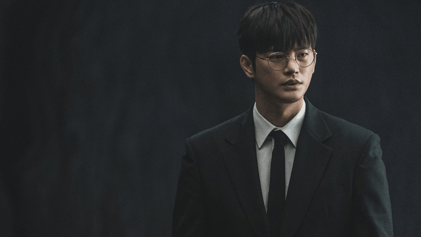 A young man in a black suit and tie stands against a dark, moody background, his serious expression highlighted by soft, dramatic lighting.