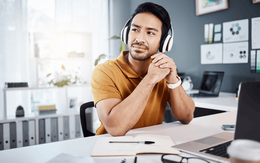 A man wearing headphones sits at a desk in a bright, organized workspace, gazing thoughtfully with a smile.
