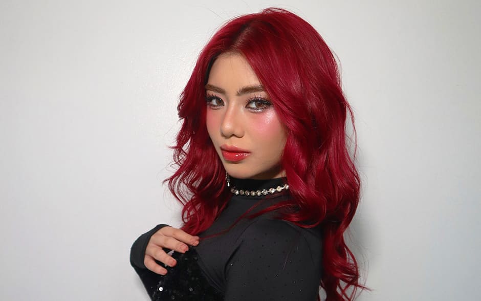 BINI Mikha striking a confident pose with vibrant red hair, wearing a black sequined top and a red skirt.