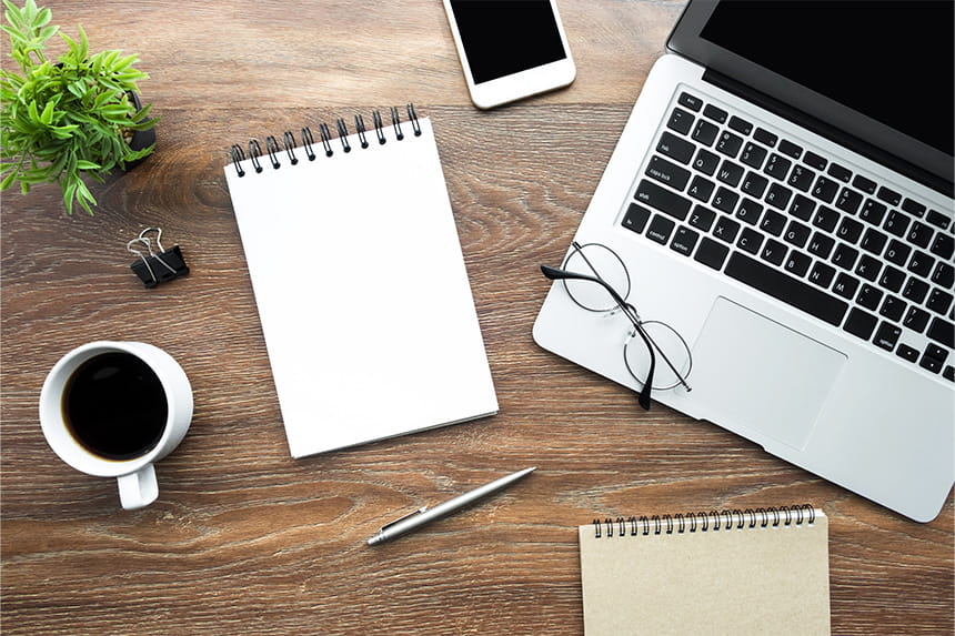 A person in a green sweater reviews a colorful planner at a desk with a laptop, digital calendar on a tablet, notebook, and succulents.