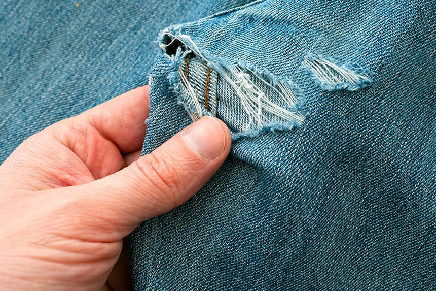 A manâs hand inspecting a hole in a pair of denim jeans.