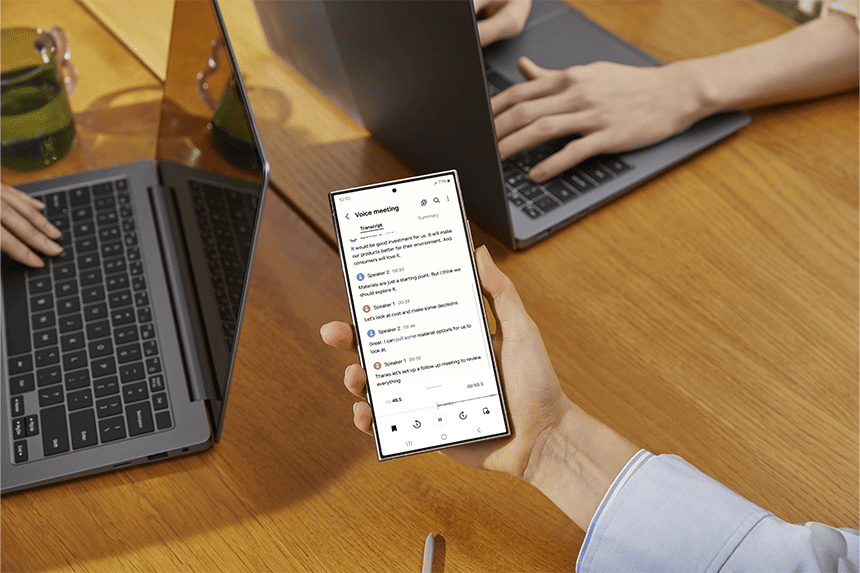 A person holding a smartphone displaying a voice meeting transcription while another person types on a laptop in the background.