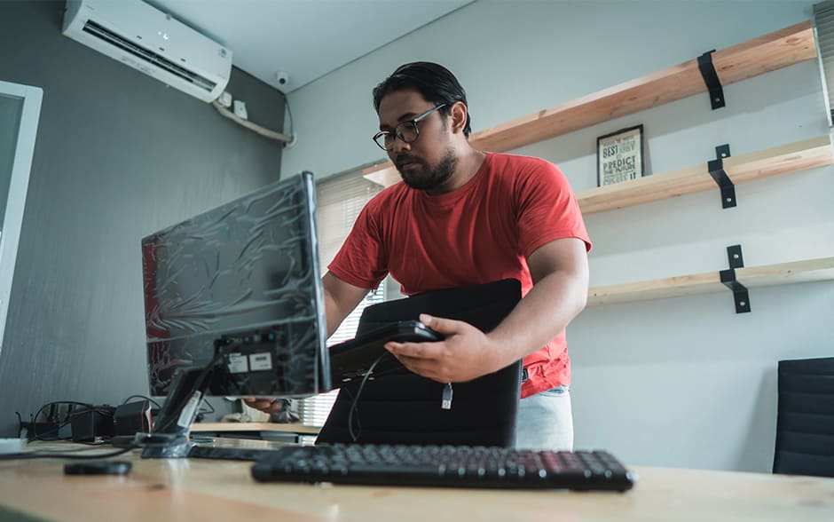 A young Asian man setting up a new monitor and keyboard on his desk.]