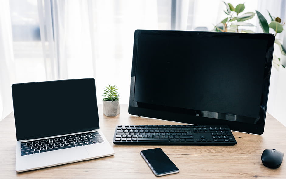 An open laptop, computer, smartphone, and potted plants on a wooden table.