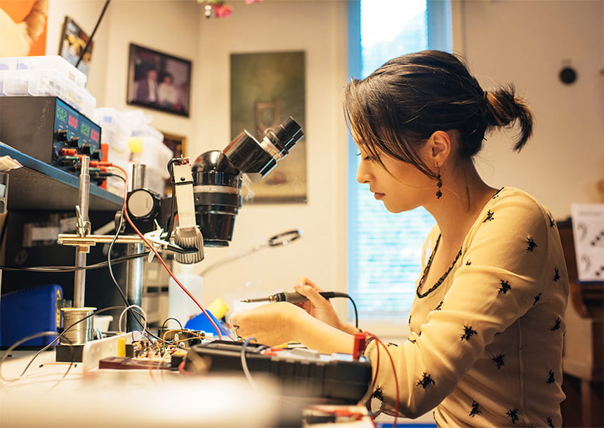 A woman soldering at an electronics lab workstation with a microscope and scattered components.