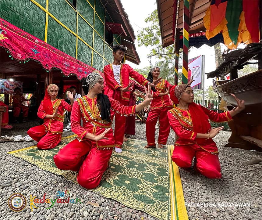 Group of dancers in red and gold attire performing on a green mat outdoors.