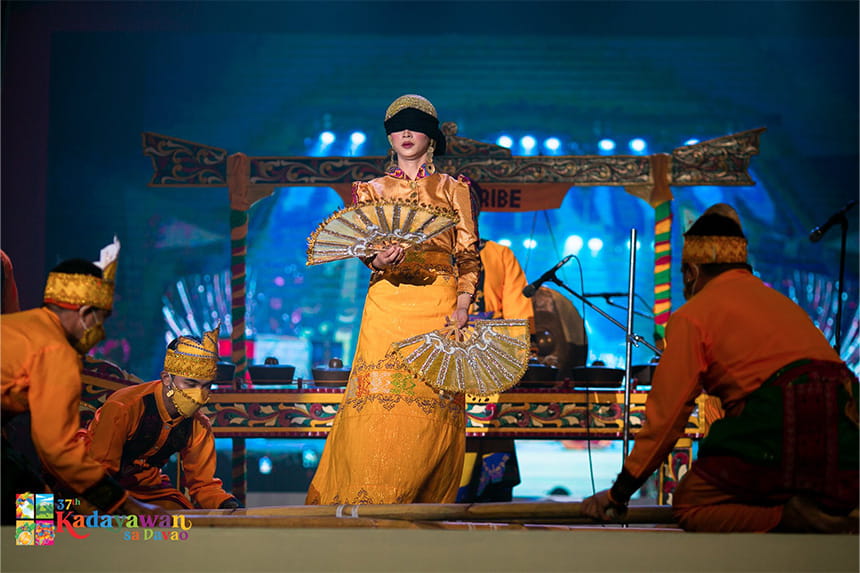 A cultural performance featuring a central figure in a golden-yellow costume with fans, surrounded by orange-clad performers on a vibrantly lit stage.