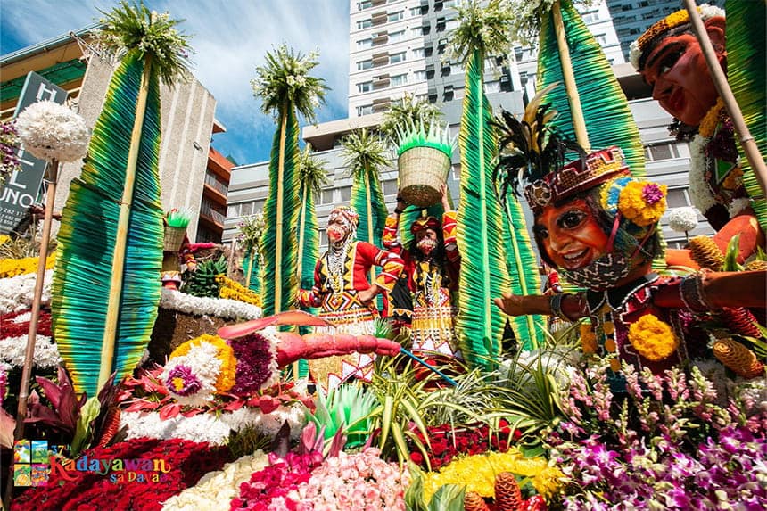 A parade float with colorful floral arrangements and people in traditional costumes, set against tall green fronds and urban buildings.