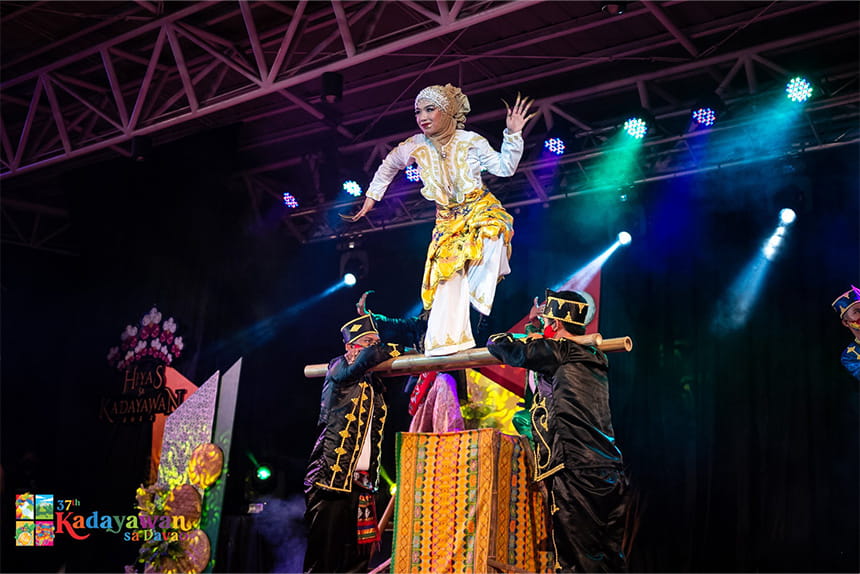 A performer balances on a bamboo pole, supported by others on stage during a cultural performance with colorful lighting.