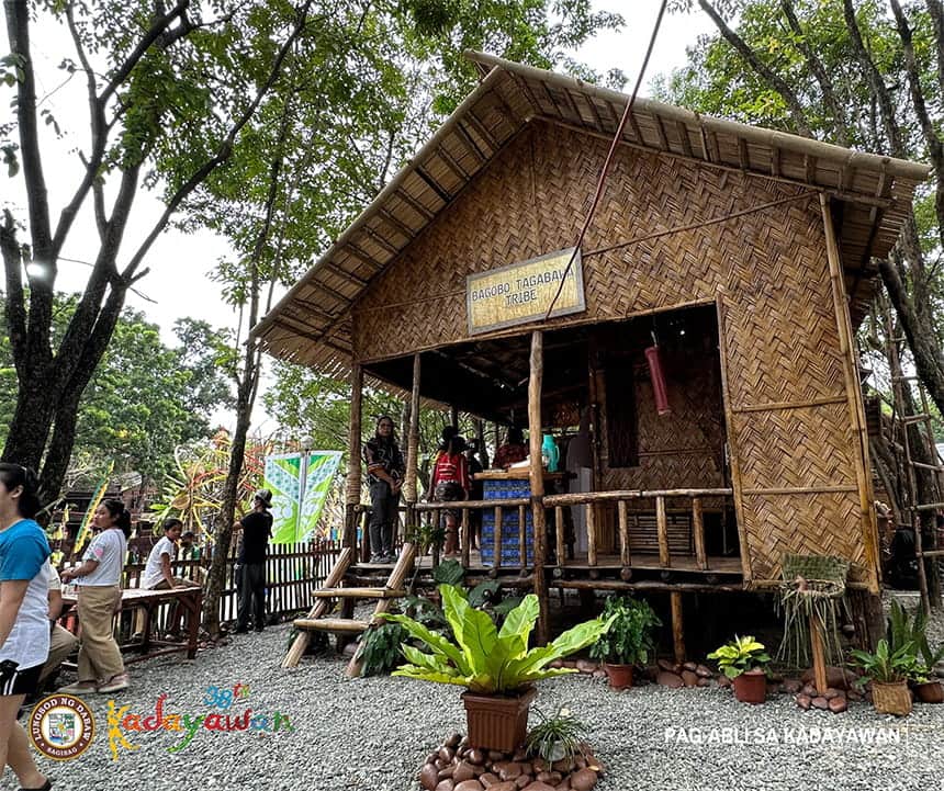 A traditional thatched wooden hut elevated on stilts with people gathered around, surrounded by trees and plants.