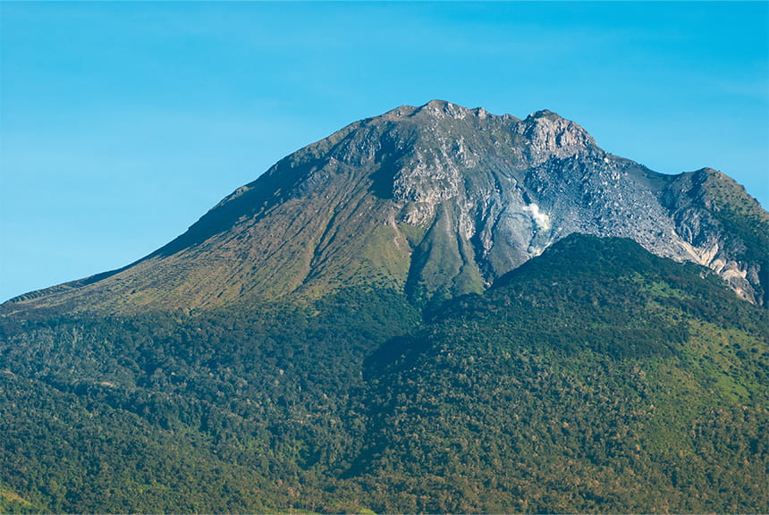 A volcanic mountain with rocky peaks and forested slopes under a blue sky.