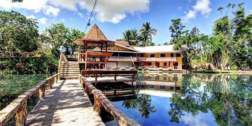 Resort with a wooden walkway leading to a pavilion over water and a two-story building surrounded by trees.