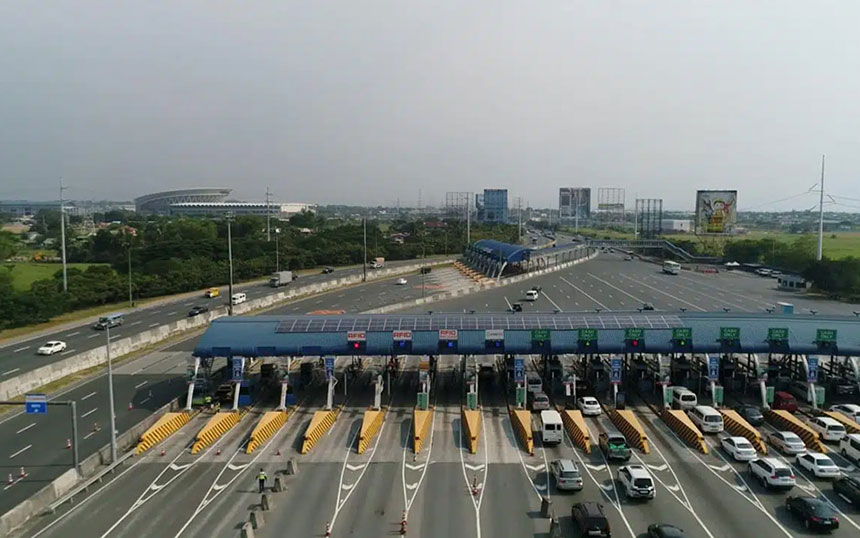 Drone view of a long, open highway flanked by lush green fields and a river, with a city skyline visible in the distance under a bright blue sky, symbolizing connectivity and progress.