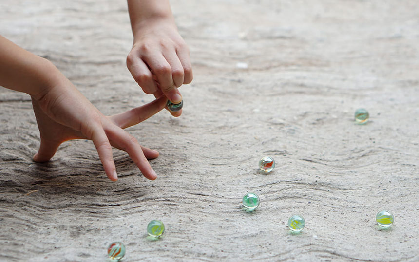 An engaging close-up of a traditional marble game in progress, showcasing the dexterous hands of a player poised to shoot a marble with precision, surrounded by scattered colorful marbles on a textured ground.