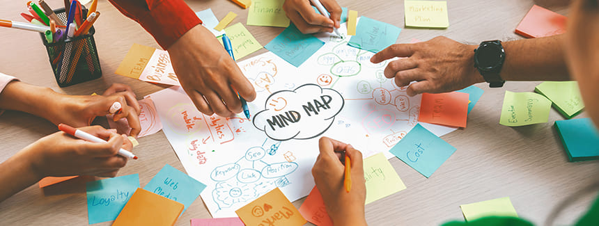 A group of people collaborating on a mind map drawn on paper surrounded by colorful sticky notes and pens on a desk.