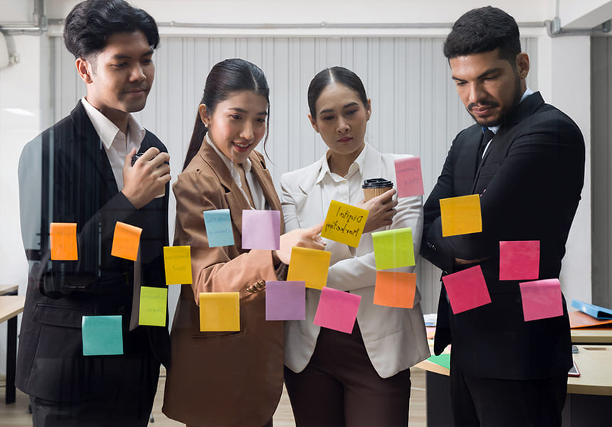 Four professionals brainstorming in front of a glass wall covered with colorful sticky notes in an office setting.