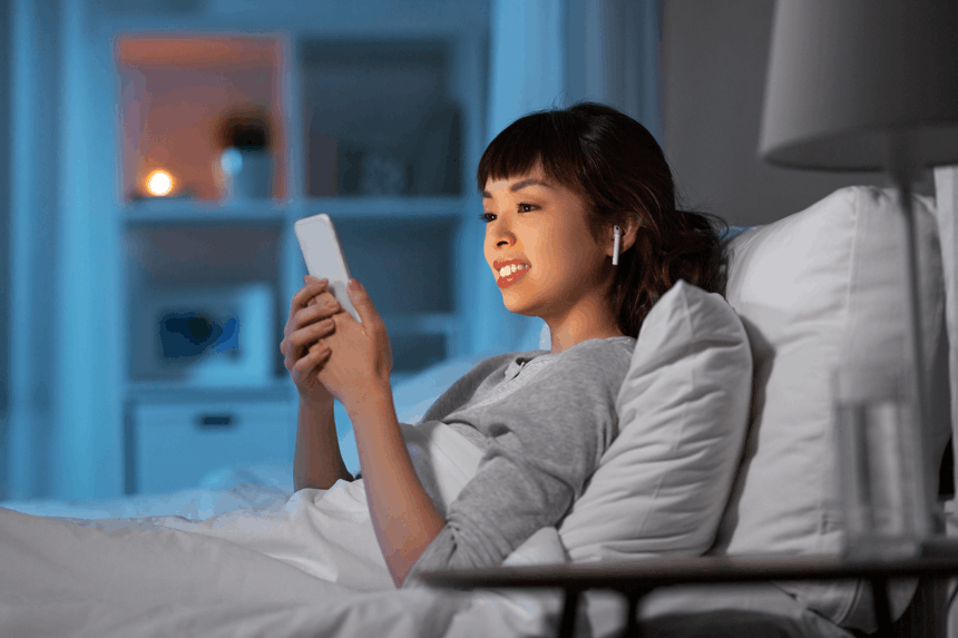 A woman in bed smiles while using her smartphone, illuminated by soft blue lighting in a cozy, dimly lit bedroom.