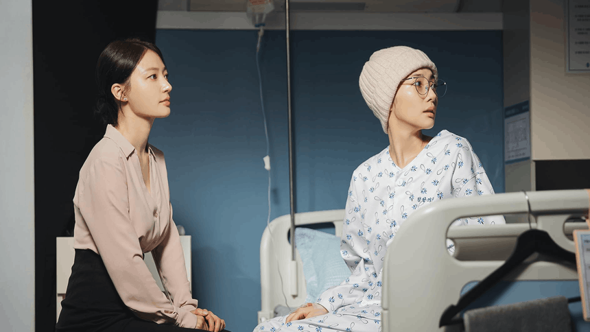 Two women sit in a hospital room; one in a patient gown and hat looks concerned, while the other listens attentively under soft, focused lighting.