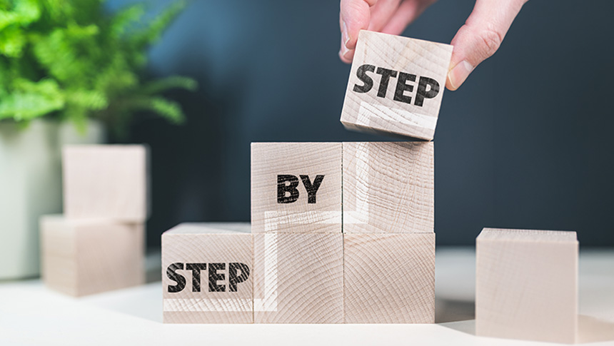 A hand stacking wooden blocks that spell out "step by step" to form a staircase.