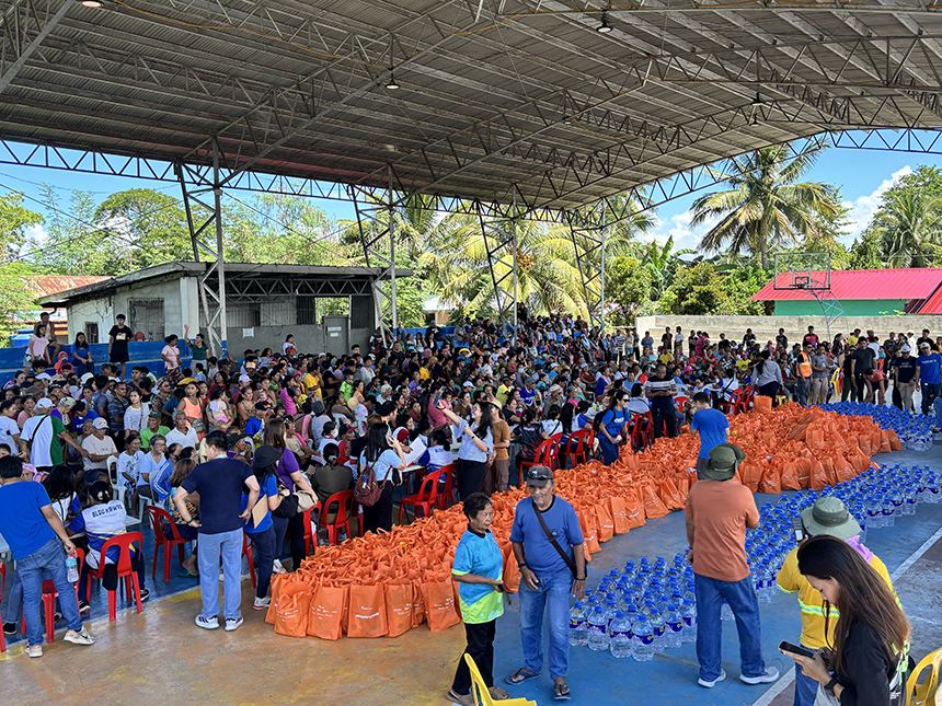 A large crowd gathers in a covered court for relief goods and bottled water distribution event.