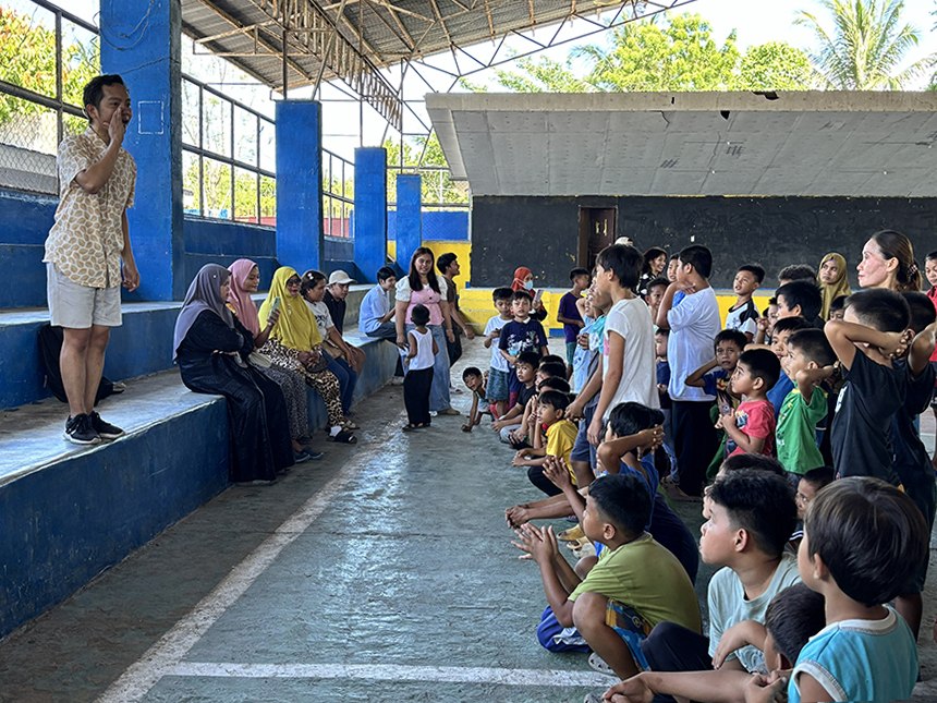A volunteer addresses children and parents during a psychosocial support session in a covered court.