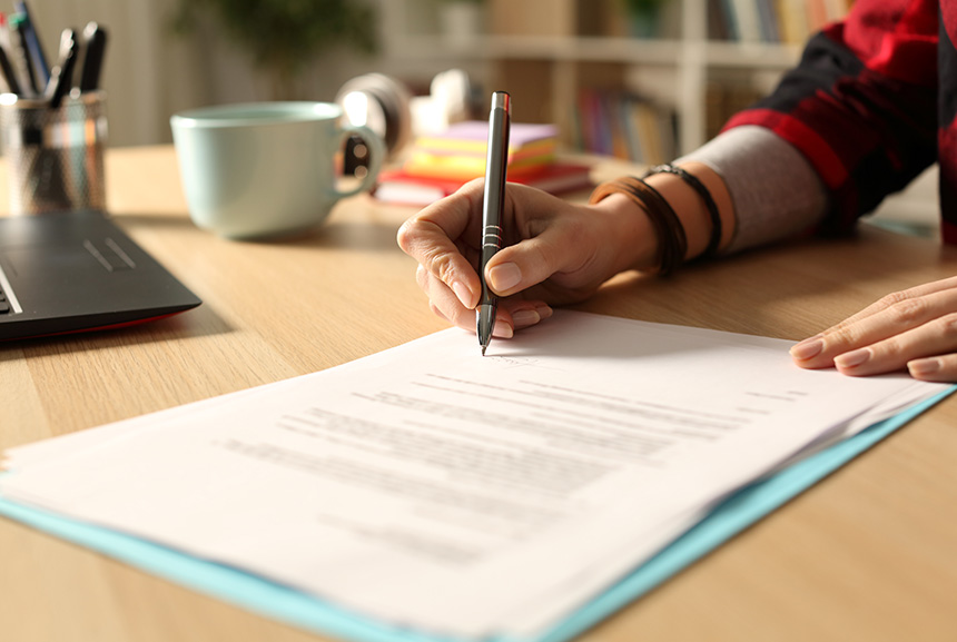 Close-up of a personâs hands writing on paper, with a laptop, cup, and stationery holder on a wooden desk.