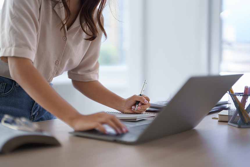A student taking notes, writing in a notebook while checking the laptop.