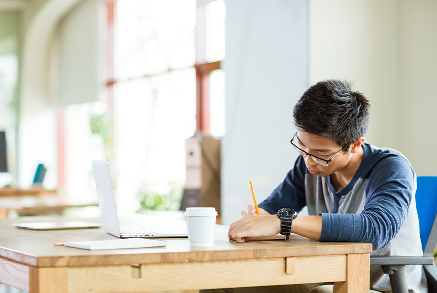 Student writing notes beside a laptop in a study area.